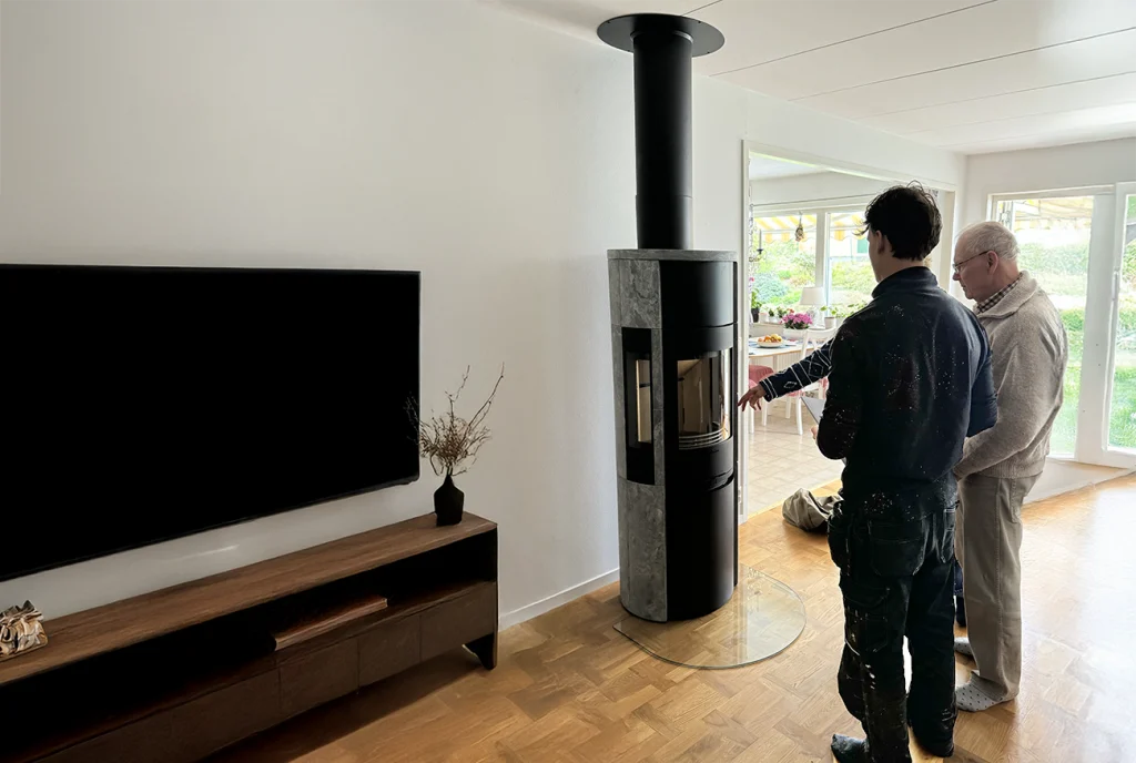 Två personer står i ett ljust vardagsrum bredvid en modern cylindrisk vedspis. Den ena personen ser ut att förklara eller demonstrera kaminen medan den andra lyssnar. På den motsatta väggen finns en TV och ett träskåp.