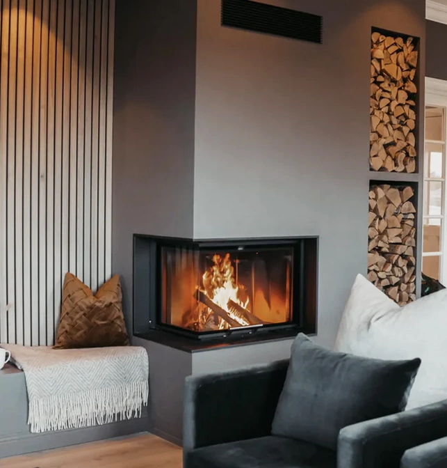 Ett modernt vardagsrum med en hörnspis som eldas med ved, en mörkgrå fåtölj, träförvaring på väggen och en mysig fönsterplats med kudde och filt.