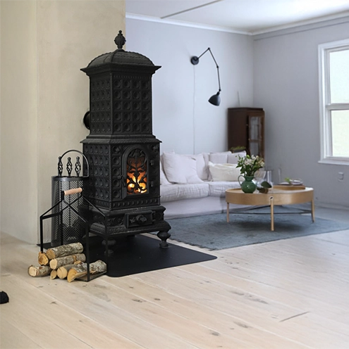 En svart gjutjärnskamin med en brasa i står bredvid en stapel ved i ett ljust, modernt vardagsrum med ljust trägolv, en vit soffa, ett runt soffbord och ett stort fönster.