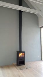 En modern svart braskamin med ett högt skorstensrör är placerad mot en ljusgrå vägg på ett ljust trägolv. Det brinner en liten brasa i kaminen och det kommer in naturligt ljus från höger.