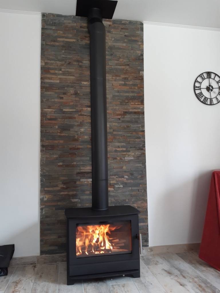 En svart vedeldad kamin med synlig eld inuti står mot en strukturerad stenvägg, med ett högt svart skorstensrör som sträcker sig uppåt. En svartvit klocka hänger på den vita väggen till höger.