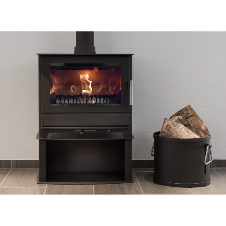 Heta Inspire 55H, en modern svart braskamin med synlig eld inuti, står på ett klinkergolv bredvid en svart metallhink med ved mot en ljusgrå vägg.