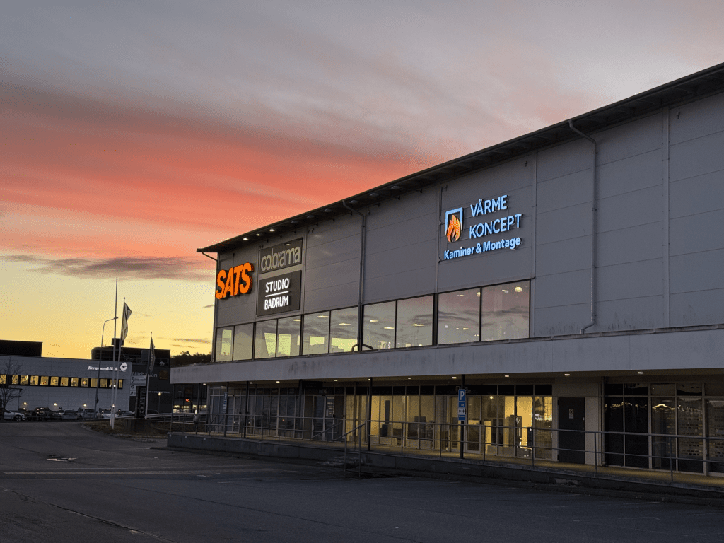 En kommersiell byggnad i solnedgången med upplysta skyltar för SATS gym, databana och Värme Koncept. Himlen är färgglad med orange och rosa nyanser. Parkeringsplatsen framför är i stort sett tom.