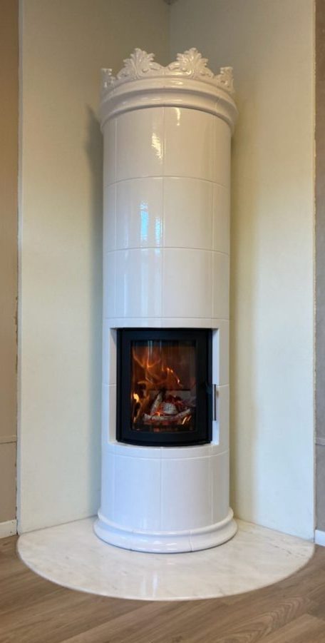 En hög, cylindrisk vit keramikspis med utsmyckade detaljer på ovansidan står i hörnet av ett rum, med en eld som brinner bakom en glasdörr på framsidan. Kaminen står på en rund, vit marmorbas.