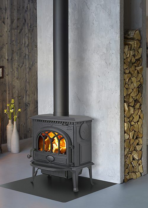 En svart vedspis av gjutjärn från Jötul med ett högt skorstensrör står på en svart matta framför en vit vägg, med ved prydligt staplad i en nisch bredvid och en vas med gula blommor i bakgrunden.