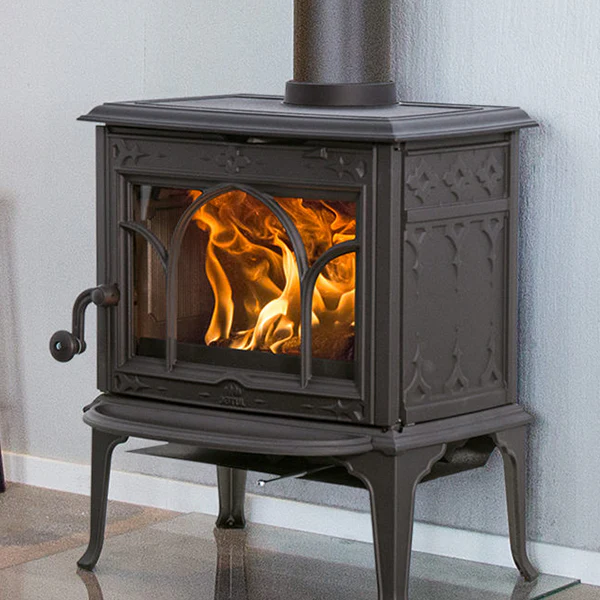 En svart vedspis i gjutjärn från Jötul med glasdörr avslöjar glödande lågor inuti och står på en reflekterande yta mot en ljus vägg.