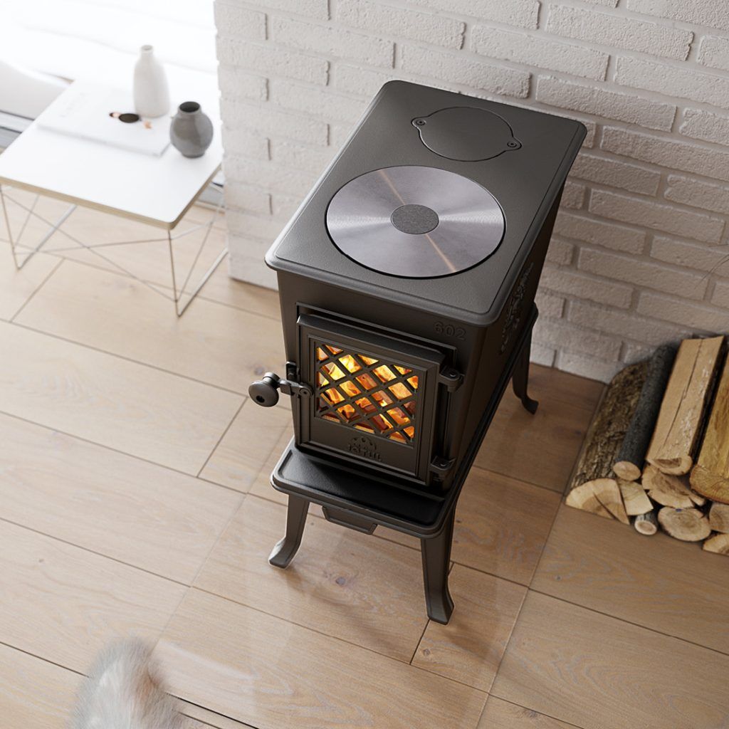 En svart Jötul-vedspis med en glasdörr som visar en eld inuti står på ett ljust trägolv bredvid staplade vedträn och en vit tegelvägg. I närheten står ett vitt sidobord med keramik.