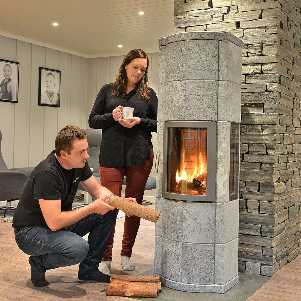 En man står på knä och lägger ved i en Norsk Kleber-stenkamin medan en kvinna står bredvid och håller i en mugg, båda i ett mysigt vardagsrum med familjefoton på väggen.