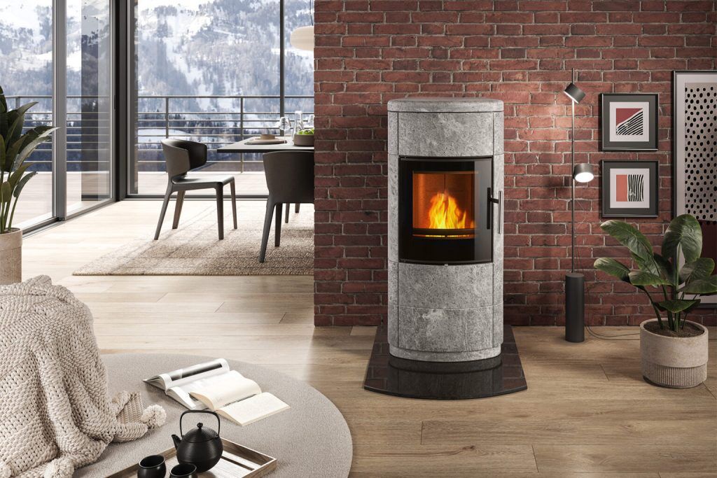 Ett mysigt, modernt vardagsrum med en Norsk Kleber stenkamin mot en tegelvägg, eld som brinner inuti, öppen bok och mugg på en matta, växter, väggkonst och en matplats med utsikt över bergen som syns genom stora fönster.