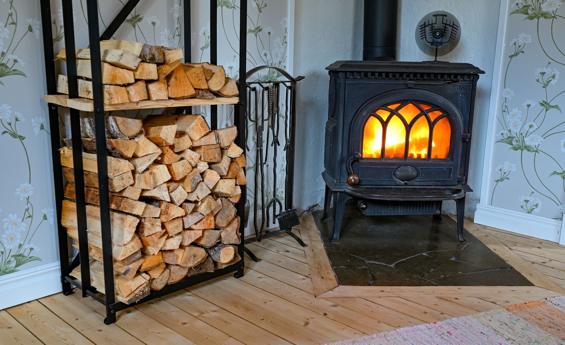 En svart vedeldad kamin, utformad efter en braskamininstallation, står på en kaklad eldstad bredvid en metallhylla med ved. Rummet har trägolv, blommig tapet och synliga eldstadsverktyg.