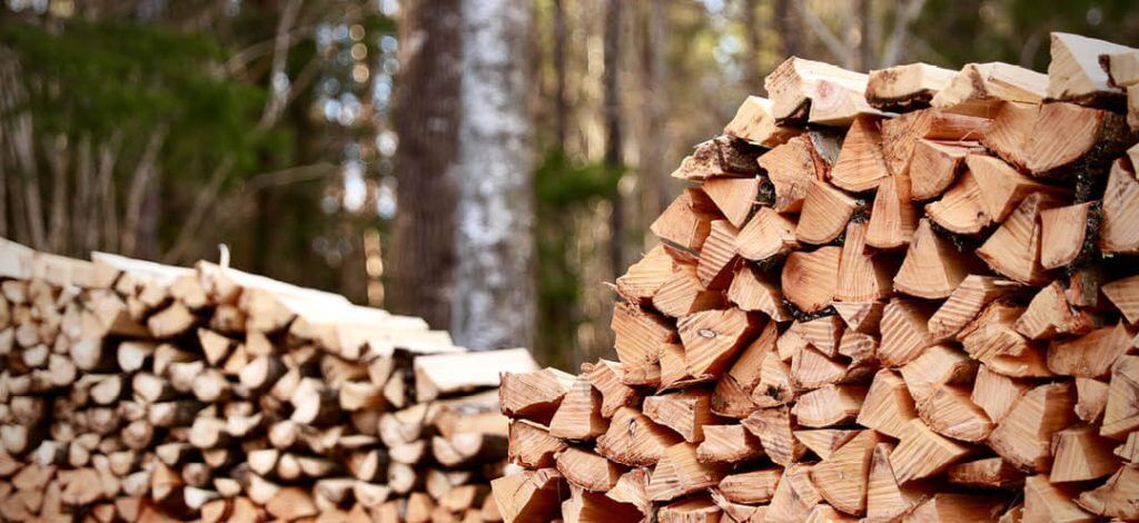 Staplar av splitved är prydligt staplade utomhus i ett skogsområde, med träd och grönska som syns i den suddiga bakgrunden.
