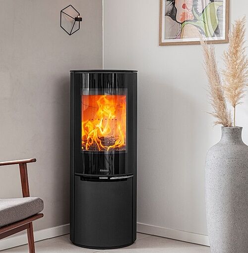 Rund Kamin: Topp 7 (Bäst i Test Design & Funktion) 2 En modern rund kamin svart vedeldad spis med en glasdörr som visar ljusa lågor är placerad i ett mysigt hörn nära en beige stol, vit vas med pampasgräs och väggkonst på en ljus vägg.