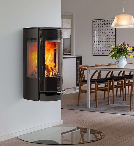 Rund Kamin: Topp 7 (Bäst i Test Design & Funktion) 4 En modern svart rund kamin väggmonterad vedspis med en eld som brinner inuti, placerad i ett minimalistiskt vardagsrum med ljusa trägolv och en eldstad i glas. I bakgrunden skymtar en matplats med träbord och stolar.