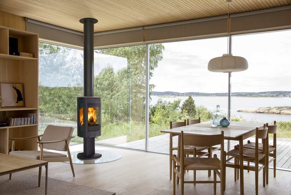Rund Kamin: Topp 7 (Bäst i Test Design & Funktion) 3 Ett modernt vardagsrum och matsal med stora fönster med utsikt över en sjö och träd, med trämöbler och en elegant rund kamin vedeldad spis med en eld inuti. Utrymmet fylls av naturligt ljus.
