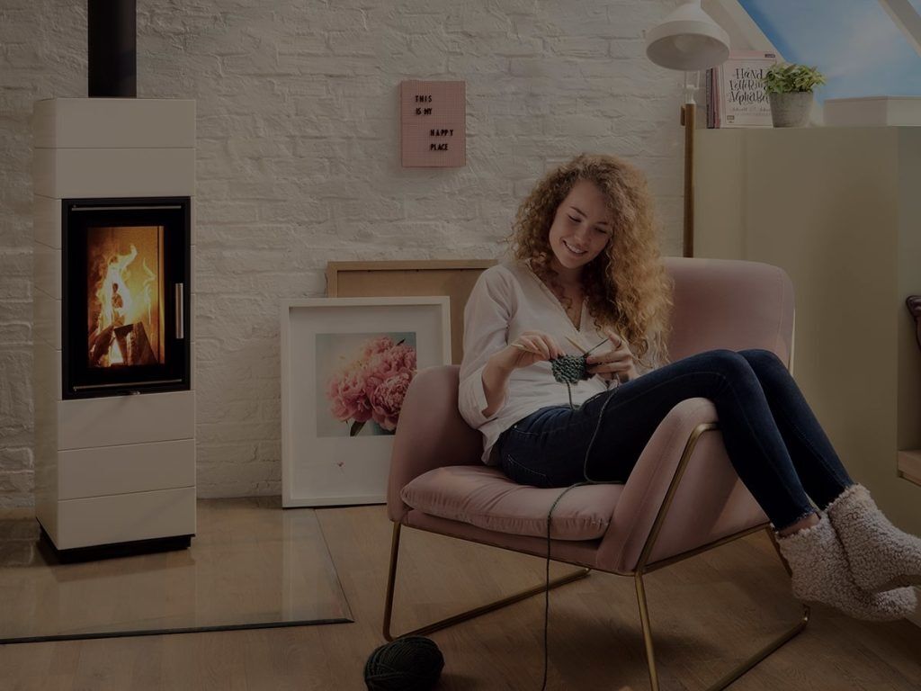 En kvinna med lockigt hår sitter i en rosa fåtölj och stickar, leende och avslappnad. Hon bär en vit topp, jeans och tofflor. Bredvid henne brinner en öppen spis och rummet är mysigt inrett med väggkonst och ett inramat blomfoto.