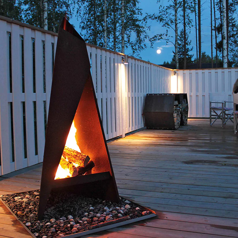 En modern triangulär utomhusspis brinner starkt på ett trädäck omgivet av ett vitt staket i skymningen, med träd och månen synlig i bakgrunden.