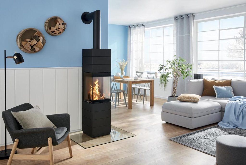 Modernt vardagsrum med en svart braskamin, mysig fåtölj, sektionssoffa, matbord och stora ljusa fönster. Veden förvaras i runda vägghyllor och en krukväxt står nära fönstret.
