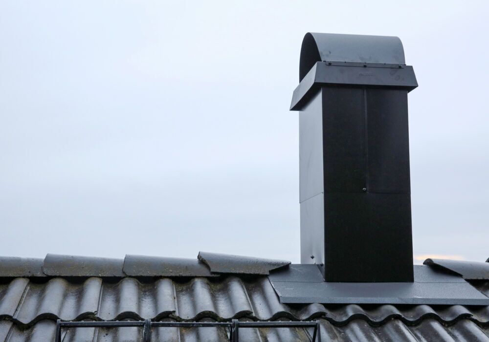 En svart metallskorsten med svängd huv monteras på ett grått tegeltak under en mulen himmel.