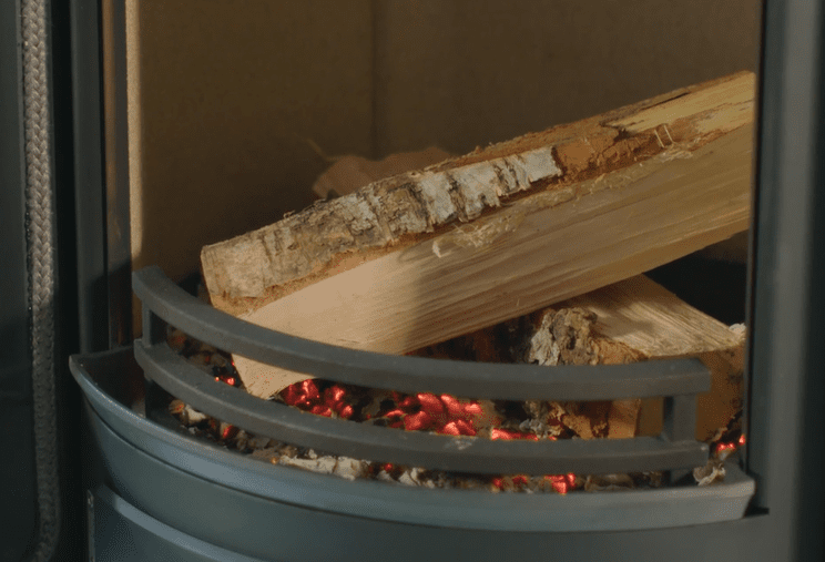 Två vedträn vilar på glödande röd glöd i en metallspis, med metallstänger framför och en brun bakgrund.