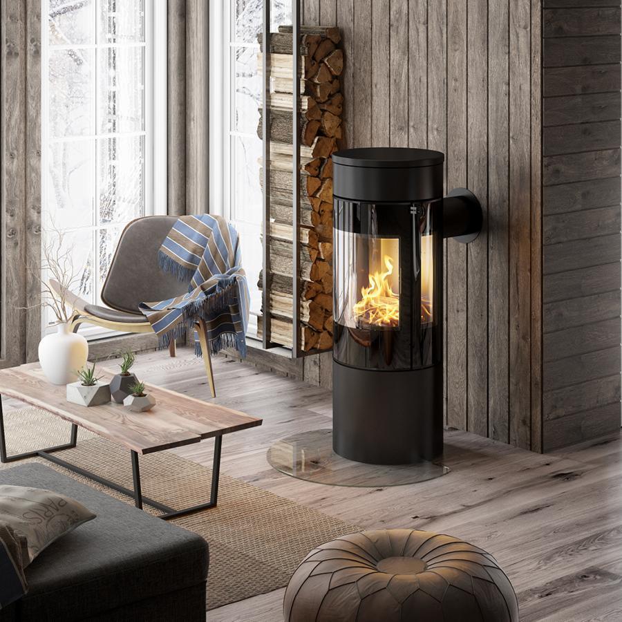 Ett mysigt modernt vardagsrum med en vedeldad kamin, en stapel ved, en stol med en blårutig filt, ett soffbord i trä och stora fönster som vetter ut mot ett snöigt landskap.