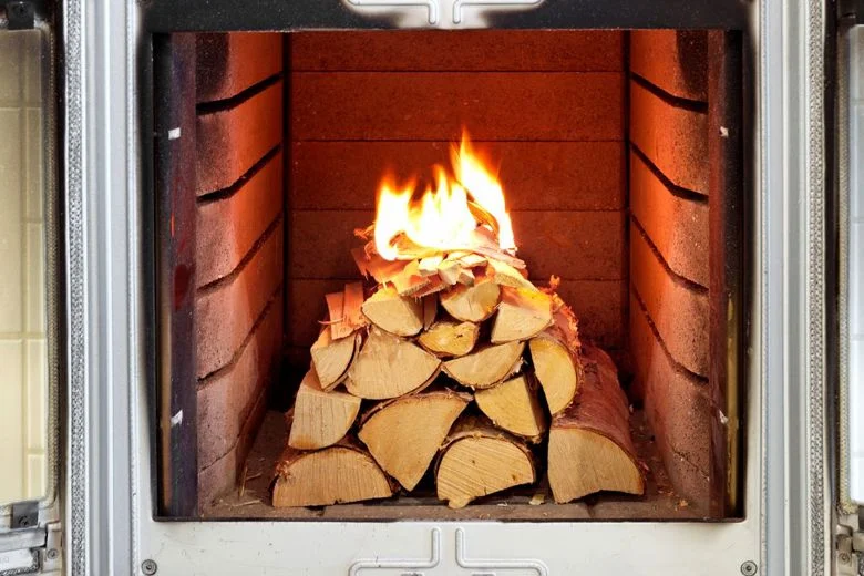 En trave ved som brinner i en modern öppen spis, med synliga lågor och glödande ved, omgiven av metall- och tegelväggar.