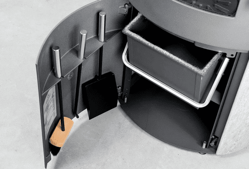 Närbild av ett svängt förvaringsutrymme med eldstadsverktyg, bl.a. borste, eldgaffel och spade, med en svart soptunna på en hylla ovanför. Facket är inbyggt i en grå enhet på ett betonggolv.