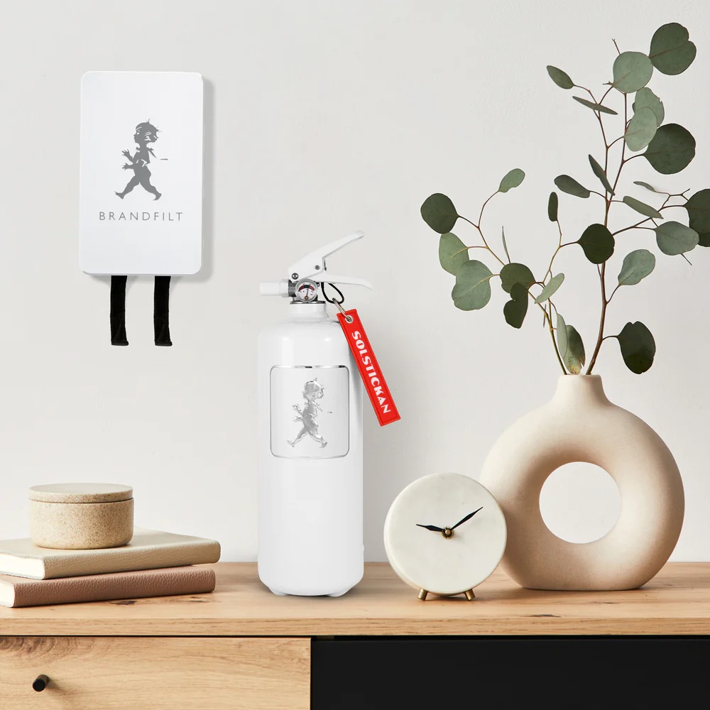 En vit brandsläckare med en röd etikett står på en träyta bredvid en rund vas med eukalyptusgrenar, en liten rund klocka och staplade lådor; en matchande väggmonterad låda hänger ovanför.