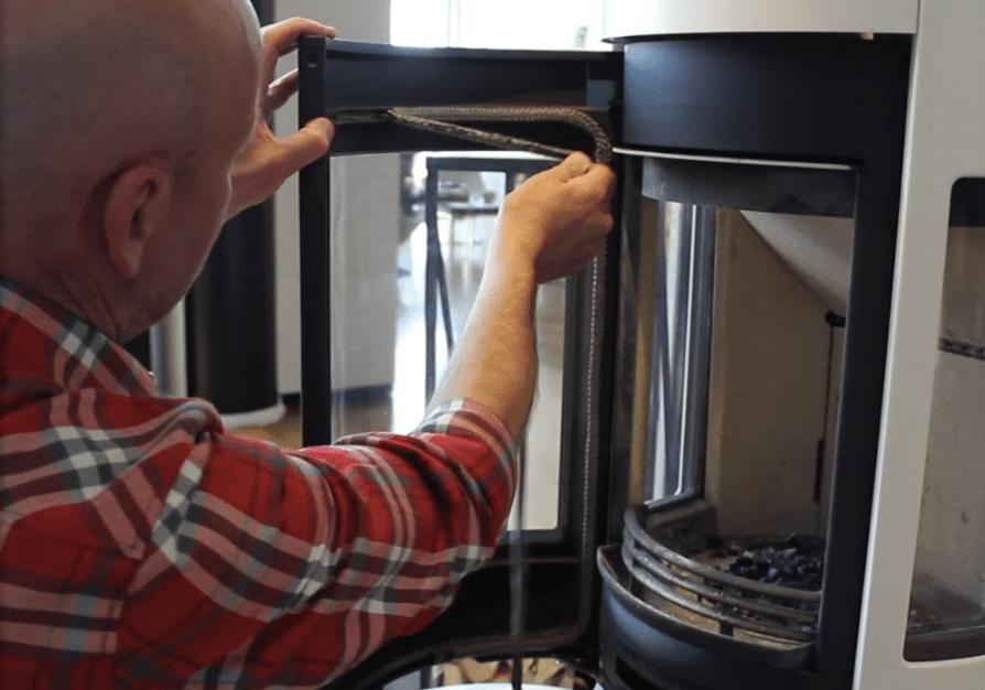 En person i rödrutig skjorta byter ut tätningen på glasdörren till en insats i en öppen spis, med dörren öppen och insidan av den öppna spisen synlig.