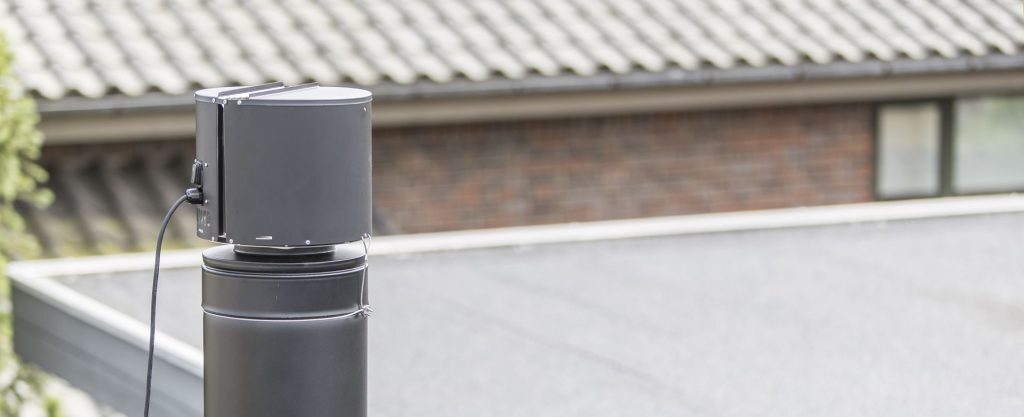 Dåligt drag i skorstenen? (Orsaker & Lösningar som fungerar) 4 Närbild av en svart cylindrisk enhet monterad ovanpå ett runt rör eller en skorsten, med en strömkabel ansluten. I bakgrunden syns ett suddigt hustak med grå bältros och ett fönster.