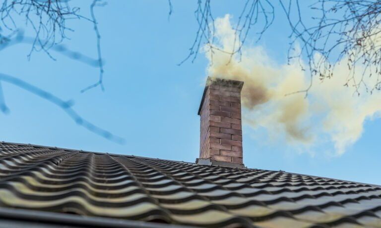 Rök stiger upp från en tegelskorsten på ett tegeltak mot en klarblå himmel, med nakna trädgrenar som ramar in scenen.