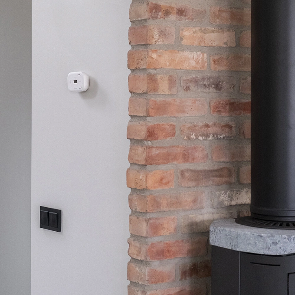 Del av innervägg med synligt rött tegel intill en vitmålad vägg. En termostat och två svarta strömbrytare är monterade på den vita väggen och till höger syns en del av en svart vedspis med stentopp.