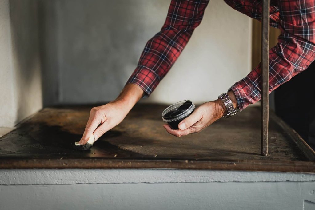 En person i rödrutig skjorta rengör en metallyta med en svamp medan han håller en rund svart behållare i andra handen. Endast armarna och händerna är synliga.