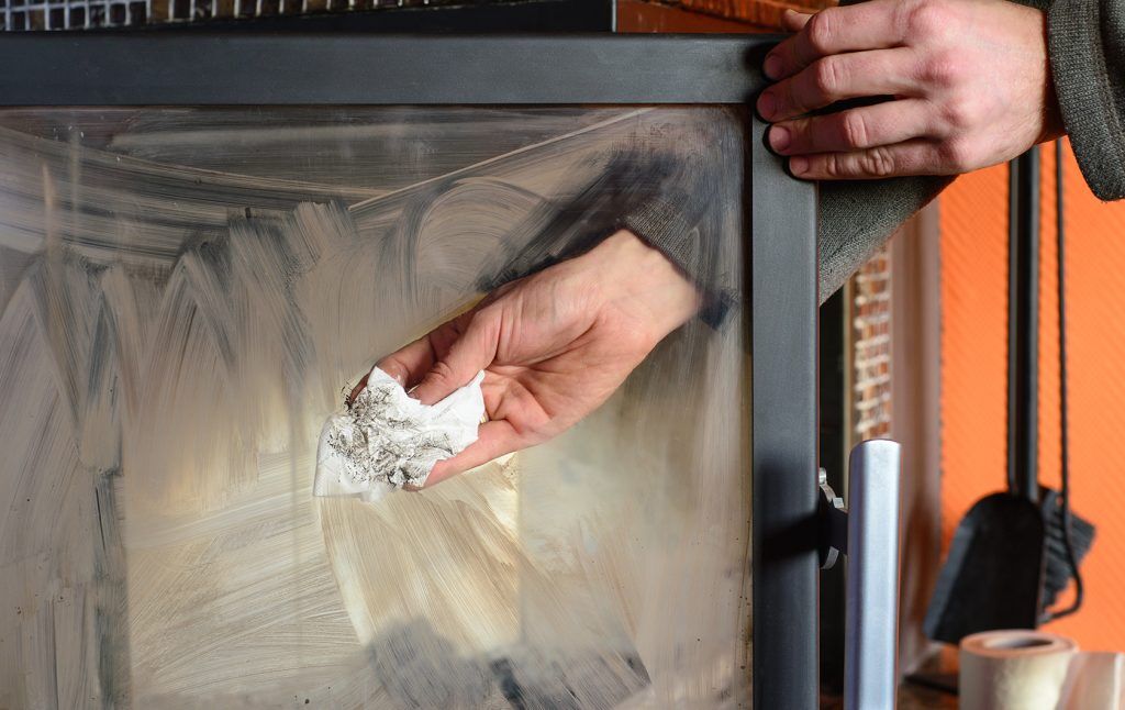 En person rengör glasdörren till en öppen spis eller vedspis och använder en pappershandduk för att torka bort sot och smuts. I bakgrunden syns eldstadsverktyg och en orange vägg.