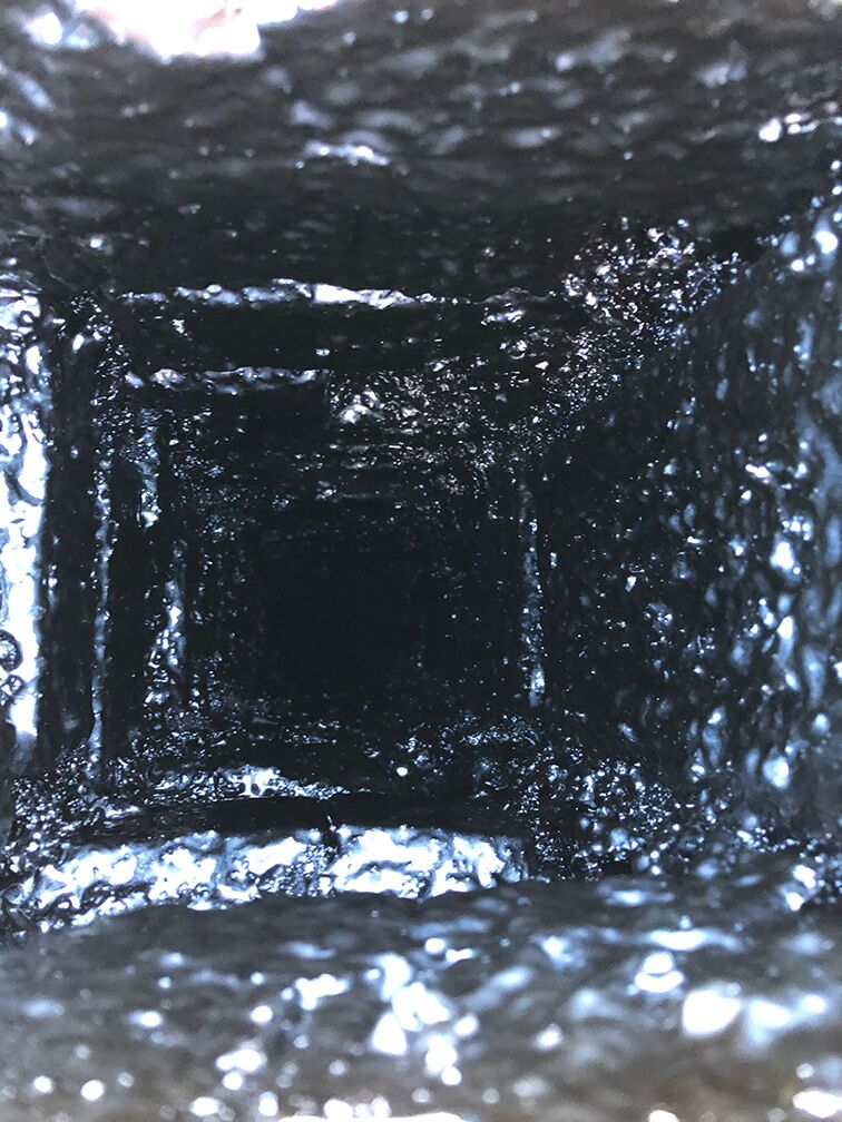 Närbild som tittar ner i en fyrkantig, sotbelagd skorsten eller ventilationsschakt med grova, blanka, svärtade väggar och ett mörkt, skuggigt centrum.