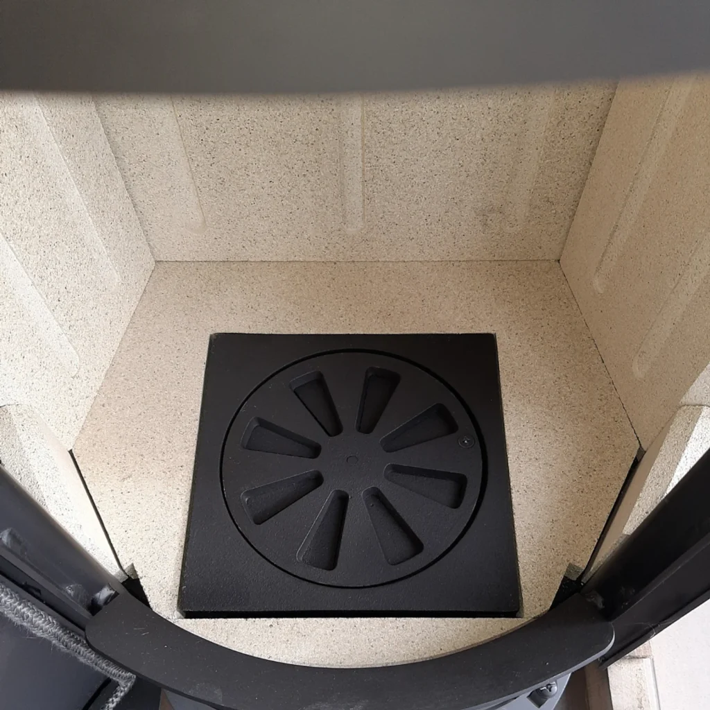 Närbild av en vedspis med ljusa tegelstenar på väggarna och ett runt svart gjutjärnsgaller med radiella spår i mitten av spisens golv.